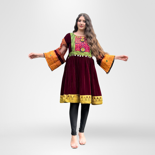 Woman wearing a burgundy velvet traditional dress with colorful embroidery in green, pink, and orange on the bodice, bell sleeves with orange and gold borders, white pom-pom trim, and gold hem detailing, Resham Dastan